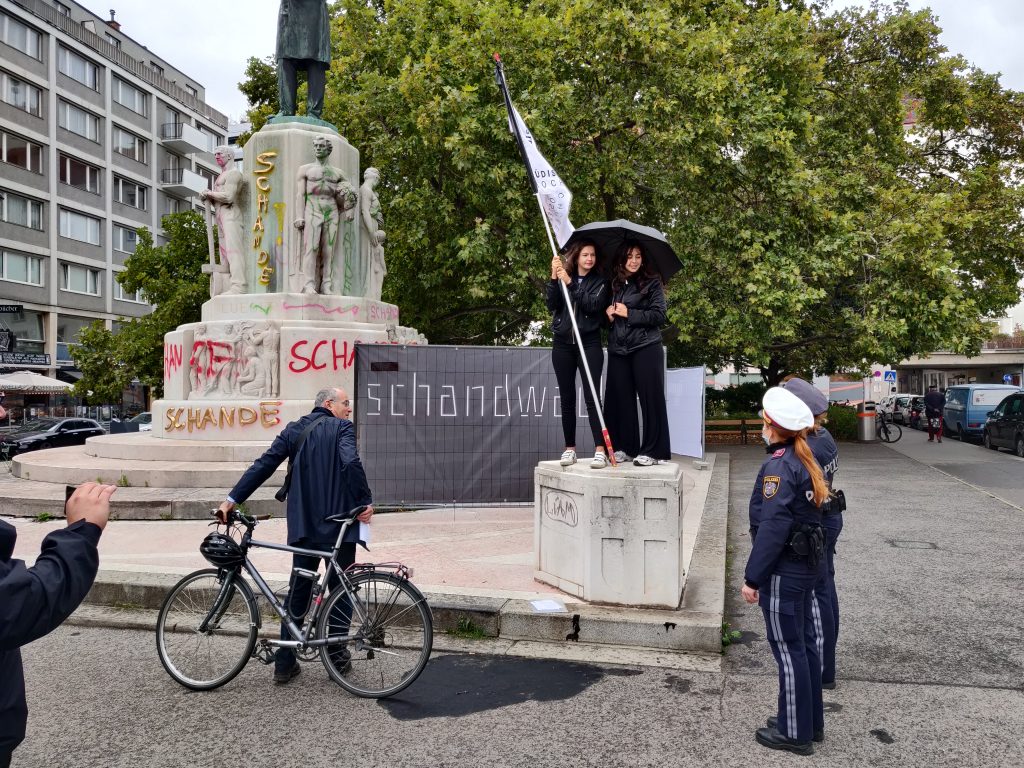Demonstrant*innen und Polizist*innen vor einem mit Graffiti besprühtem Denkmal.