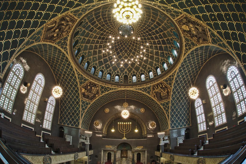 Blick in die Augsburger Synagoge: Mit grünen und goldenen Mosaiken verzierter Kuppelbau von innen, vorne auf einer Empore ein siebenarmiger Leuchter.