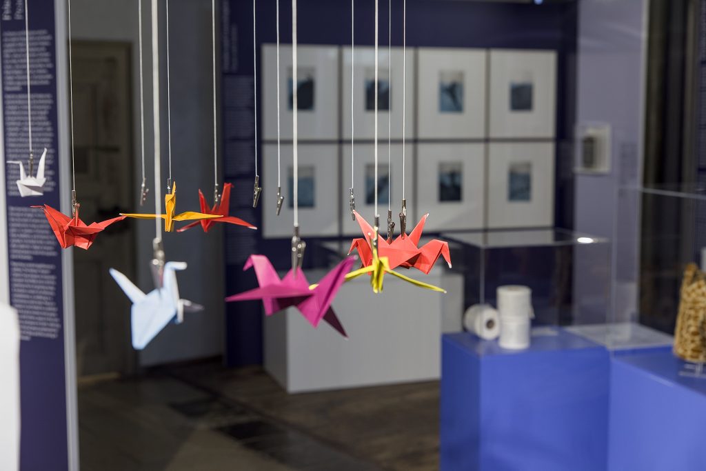 Bunte gefaltete Papier-Kraniche an Fäden hängend, im Hintergrund Vitrinen und gerahmte Kunstwerke