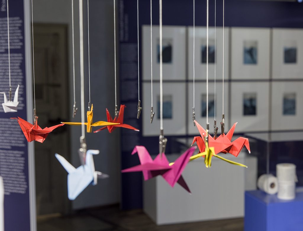 Bunte gefaltete Papier-Kraniche an Fäden hängend, im Hintergrund Vitrinen und gerahmte Kunstwerke