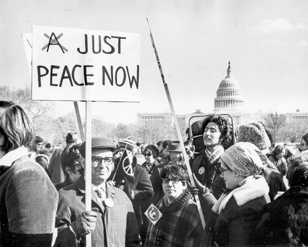 Schwarz-weiß Foto einer Demo, vorne ein Mann mit Hut und einem Plakat mit der Aufschrift "Just Peace now".