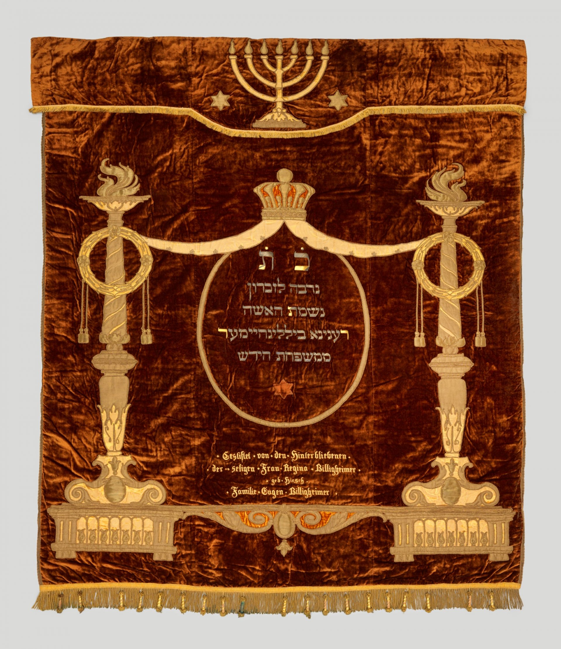 Rot-brauner Vorhang, darauf in Goldstickerei Verzierungen zum Beispiel oben einen siebenarmigen Leuchter, mittig gestickte Inschriften in Deutsch und Hebräisch.