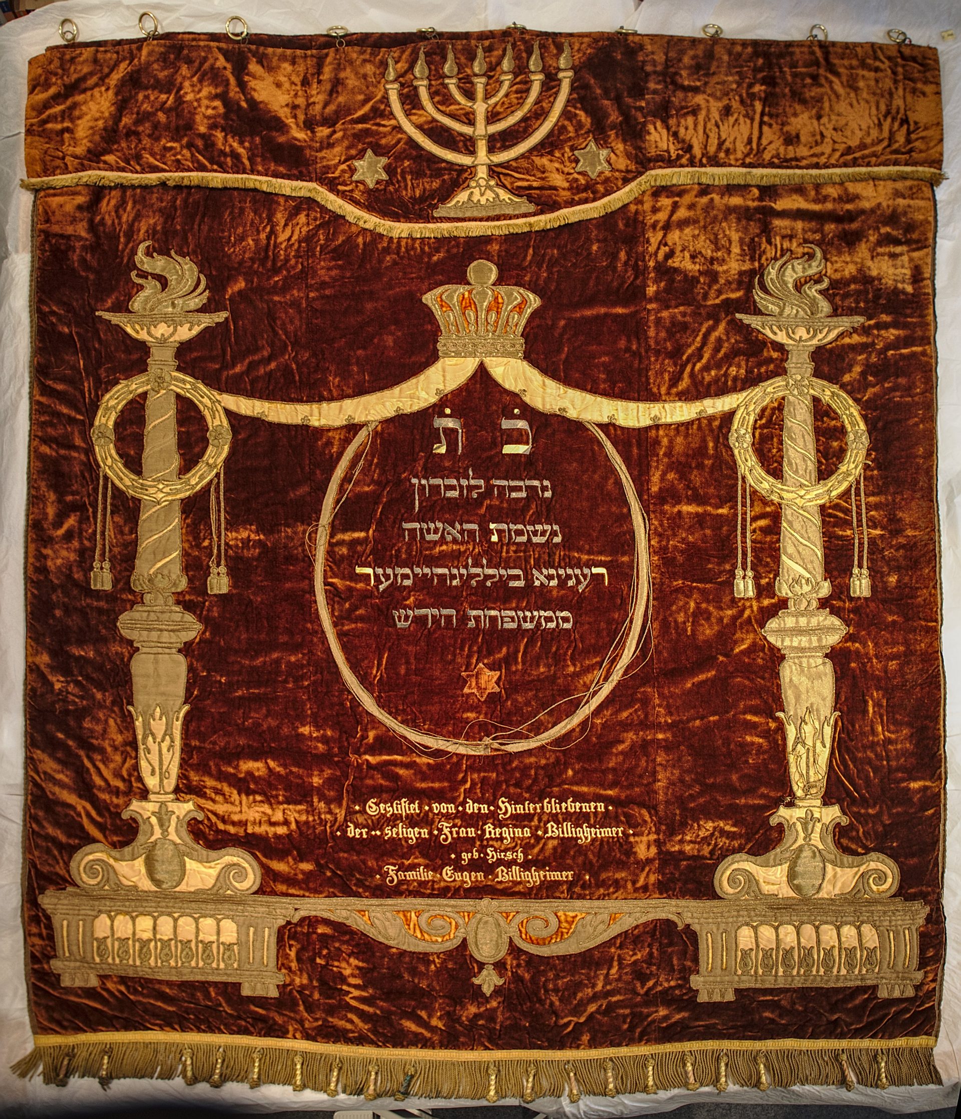 Rot-brauner Vorhang, darauf in Goldstickerei Verzierungen zum Beispiel oben einen siebenarmigen Leuchter, mittig gestickte Inschriften in Deutsch und Hebräisch.