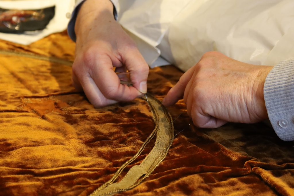 Hände mit einer Nadel, die goldgelbe Kordeln an einen rotbraunen Samststoff annähen.