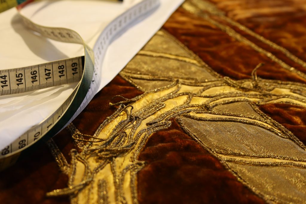 Auf dem Bild ist ein Teil eines Tora-Vorhangs zu sehen, gefertigt aus dunkelbraunem, weich glänzendem Samt. Darauf liegen goldene Stoffapplikationen, deren Konturen aus gedrehten, metallic schimmernden Kordeln bestehen. Einige dieser Kordeln haben sich gelöst und liegen ungeordnet über den Motiven. Ein weißes Maßband schlängelt sich über den Stoff – ein Hinweis auf die laufenden Restaurierungsarbeiten. Die Szene wirkt ruhig und konzentriert, als würde gerade an der Vorbereitung zur Fixierung der losen Kordeln gearbeitet.