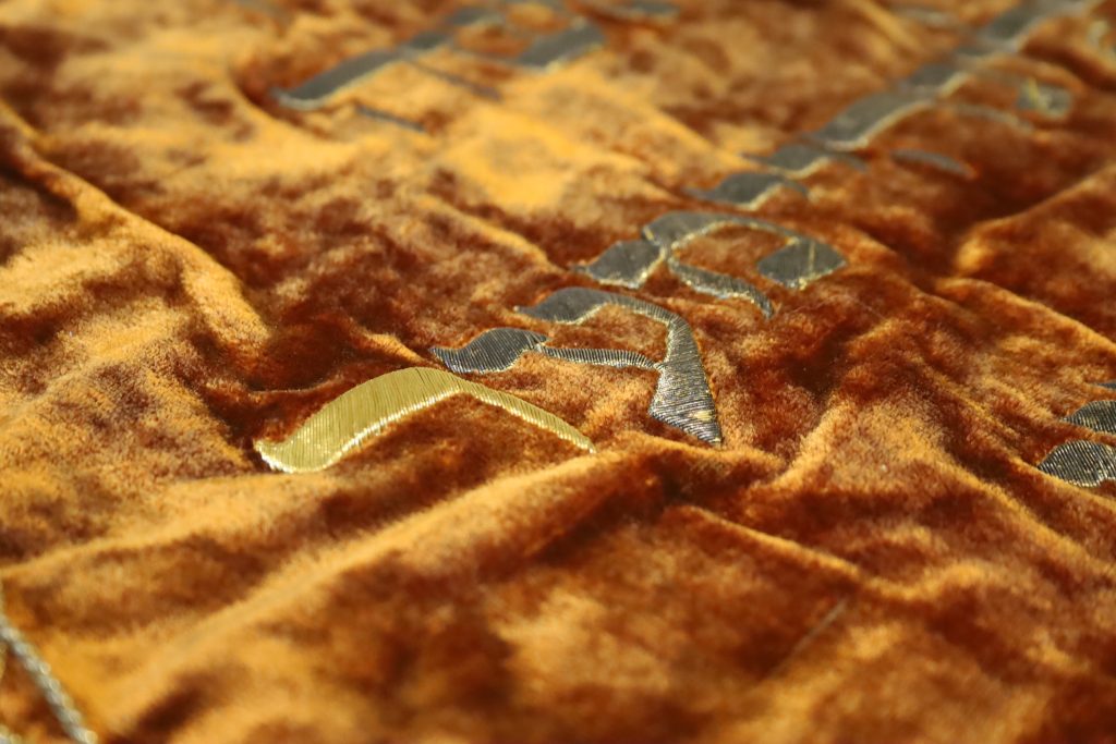 Detailaufnahme von rotbraunem Samt, darauf mit goldenem Garn gestickte hebräische Schrift