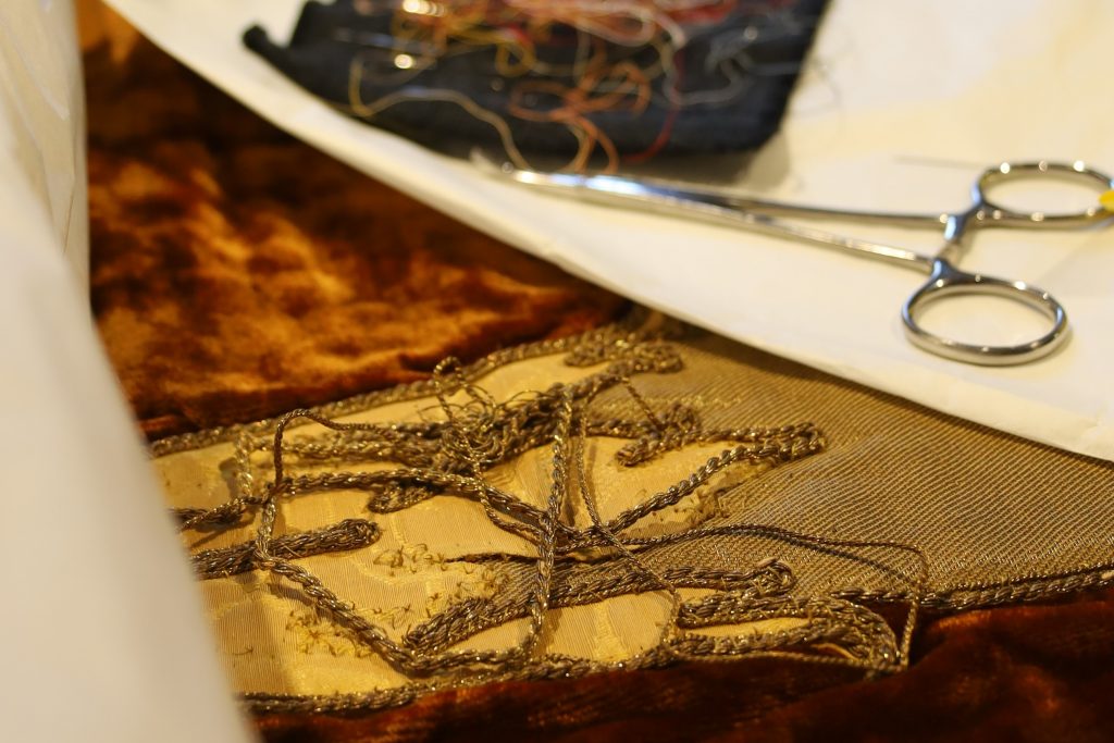 Rotbrauner Samtstoff und goldgelbe Kordeln, die offenbar nicht mehr angenäht sind. Danaben eine silberne Schere.