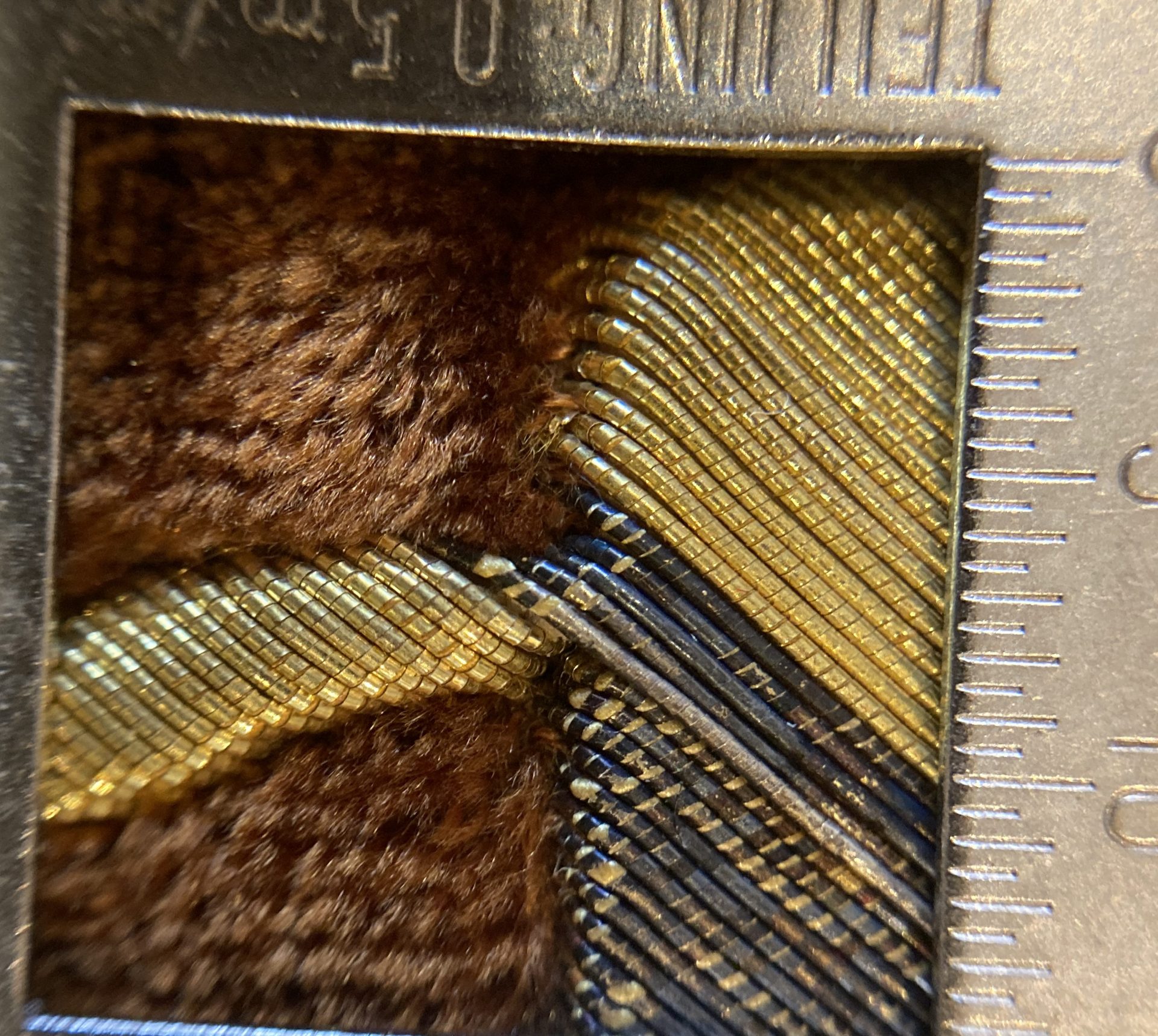 Im Bild sieht man durch ein metallisches Vergrößerungsglas auf einen kleinen Ausschnitt eines kunstvoll gearbeiteten Tora-Vorhangs. Das Sichtfenster des Instruments ist quadratisch und wird von einer Skala mit feinen Strichen und Zahlen eingerahmt. Dahinter erscheint der braun-rote Seidensamt als weiche, leicht schimmernde Fläche. Darauf liegen mehrere dicht gewundene Metallfäden, die in geschwungenen Linien verlaufen. Die Fäden glänzen teils goldfarben, teils dunkel, fast schwarz – ein Hinweis auf die Oxidation des ehemals versilberten Drahtes. Durch die starke Vergrößerung erkennt man sehr deutlich die Struktur der Metallgarnstickerei: Jeder Faden besteht aus winzigen, eng aneinanderliegenden Windungen, die wie kleine Ringe oder Schuppen aufgereiht sind. Die Oberflächen reflektieren das Licht und erzeugen ein feines Spiel aus Gold-, Braun- und dunklen Metalltönen.