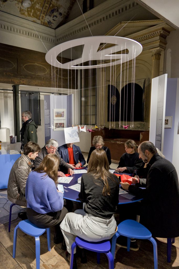 Menschen im Kultraum der ehemaligen Synagoge Kriegshaber an einem blauen Tisch, darüber ein weißes Friedenszeichen.