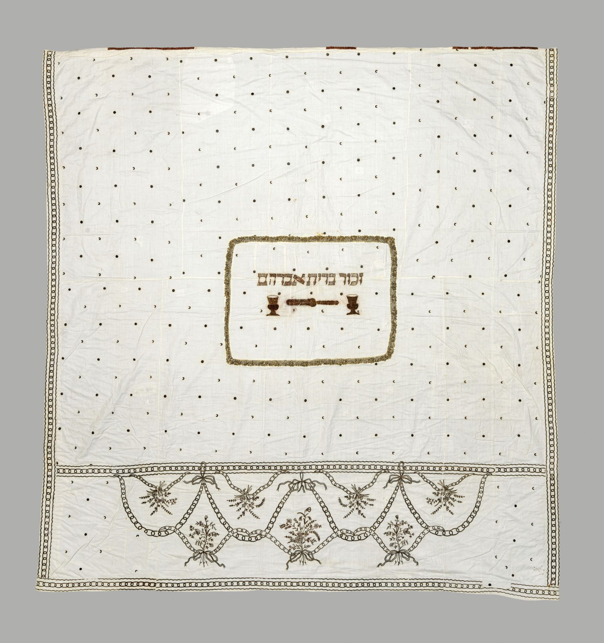 Weißer Stoffvorhang, mit Stickereien von Blumen, Ornamenten und hebräischen Buchstaben verziert.