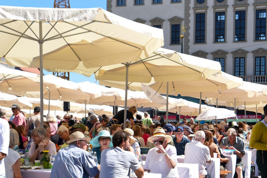 Viele Menschen sitzen zusammen an Biertischen unter großen weißen Sonnenschirmen. Im Hintergrund ist ein großes Gebäude erkennbar.