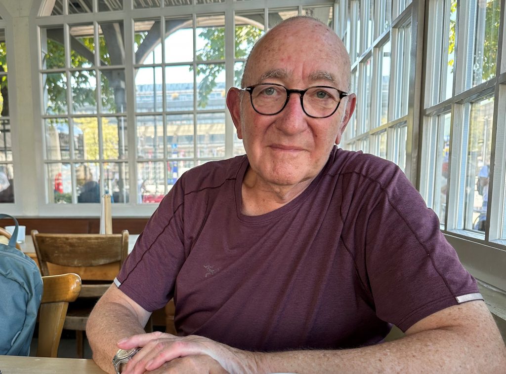 Porträt Michael Guggenheimer, ein älterer Mann sitzt an einem Tisch und schaut in die Kamera