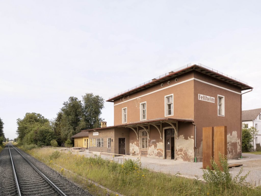 Im Vordergrund Bahnschienen, dahinter ein rötliches zweistöckiges Gebäude.